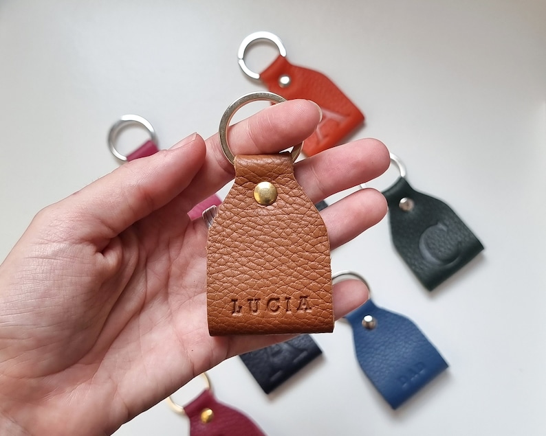 In one hand is a small leather keychain engraved with five initials (name of Lucia). Brown color, full grain leather. The keychain is on one hand and behind it you can see more keychains of other colors.