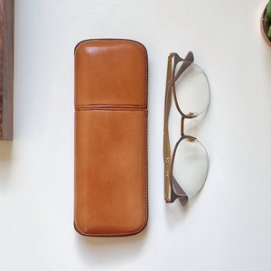 smooth tan leather eyeglasses case in  on a white table. Shown closed with eyeglasses next to it to show the proportion between the eyeglasses and the leather case.