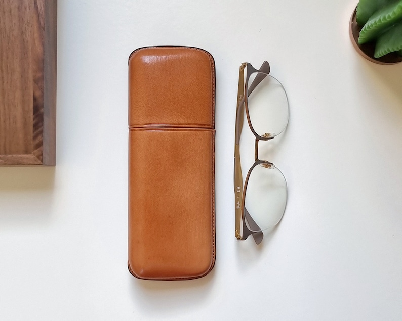 smooth tan leather eyeglasses case in  on a white table. Shown closed with eyeglasses next to it to show the proportion between the eyeglasses and the leather case.