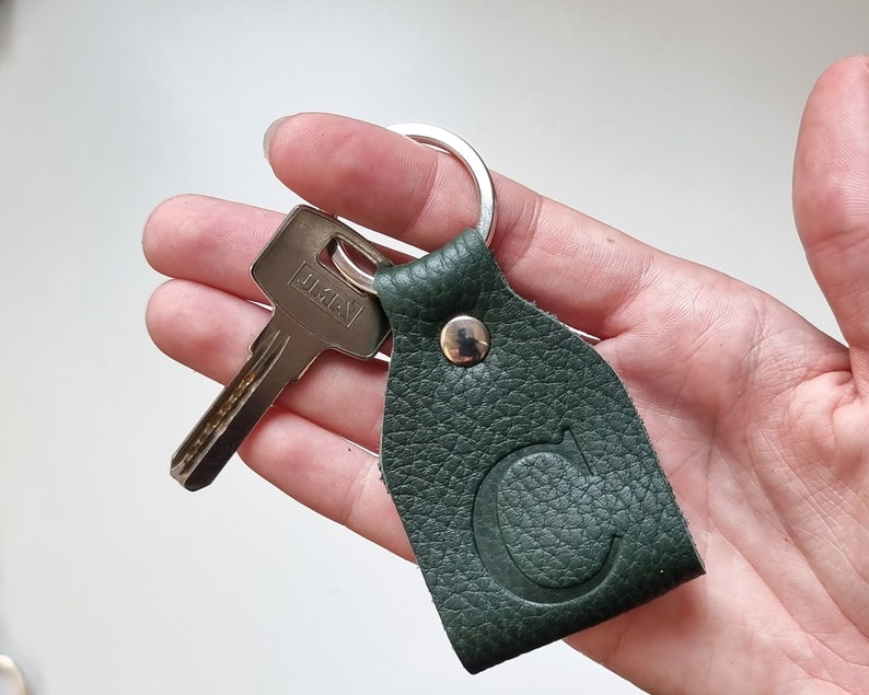 in one hand is a small key ring of soft full-grain leather in dark green engraved with the capital letter D.