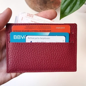 Minimalist leather card holder in burgundy color, with two card slots on each side and a central compartment where you can carry tickets or bills. In the two front slots you can see two cards and in the central compartment a ticket that comes out.