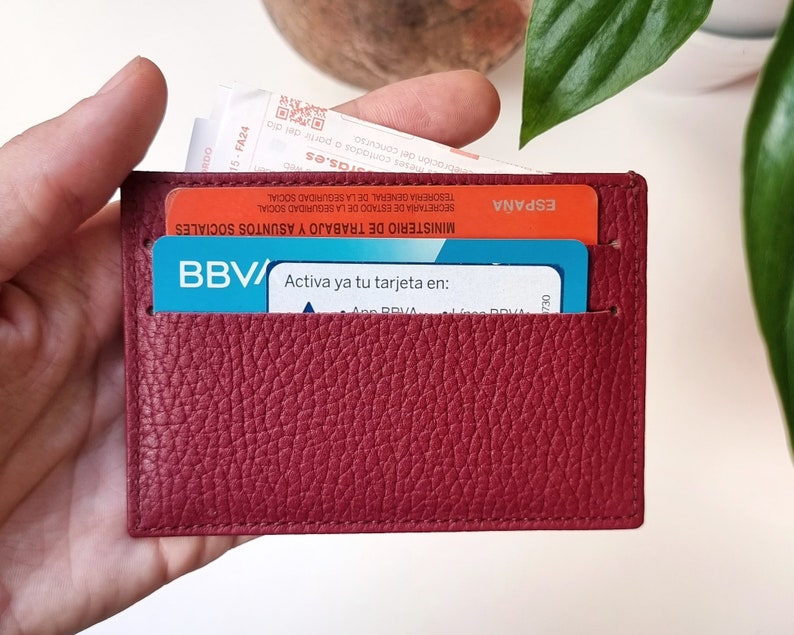 Minimalist leather card holder in burgundy color, with two card slots on each side and a central compartment where you can carry tickets or bills. In the two front slots you can see two cards and in the central compartment a ticket that comes out.