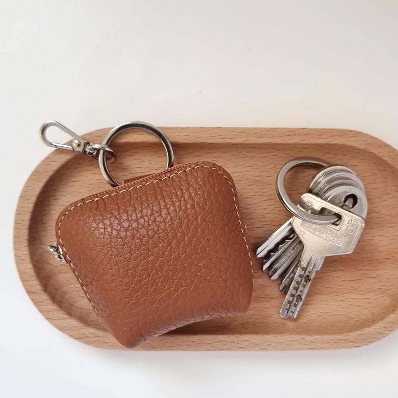 May include: A brown leather key pouch with a zipper and a silver key ring attached. Next to it, a set of keys on a silver ring rests on a wooden oval tray. The pouch has white stitching.