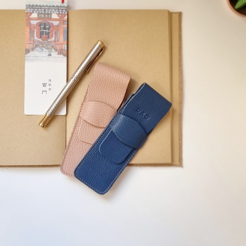 two leather pen holders with capacity for two pens or pencils each. One of the pen cases is blue and has the word DAD engraved on the flap closure. The other leather pen pouch is light pink.