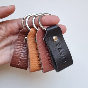 Könnte beinhalten: Vier Leder-Schlüsselanhänger mit Metallringen. Die Schlüsselanhänger sind braun, beige, rot und schwarz. Jeder Schlüsselanhänger hat einen eingravierten Namen: Daniel, Sarah, Oliver und David.