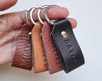 Könnte beinhalten: Vier Leder-Schlüsselanhänger mit Metallringen. Die Schlüsselanhänger sind braun, beige, rot und schwarz. Jeder Schlüsselanhänger hat einen eingravierten Namen: Daniel, Sarah, Oliver und David.