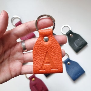 on one hand is a small key ring made of soft full grain leather in orange engraved with the capital letter A.