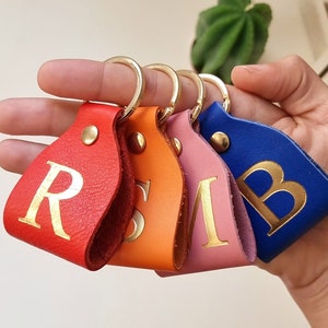 Pode incluir: Quatro chaveiros de couro com argolas de metal dourado e letras douradas. Os chaveiros são vermelho, laranja, rosa e azul. As letras nos chaveiros são R, S, M e B.