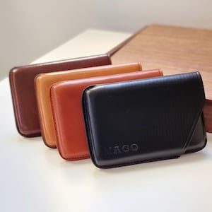 以下が含まれることがあります： 異なる色の革製のカードホルダーが4つあります。カードホルダーは左から右に、茶色、ベージュ、オレンジ、黒の順に並んでいます。黒いカードホルダーには、前面に「IAGO」という文字が刻印されています。