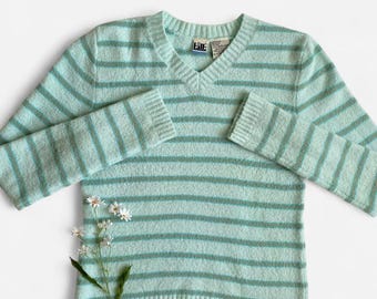 May include: A light blue V-neck sweater with horizontal stripes in a darker shade of blue-green. The sweater has long sleeves and ribbed cuffs and hem. A sprig of white flowers with yellow centers and green leaves is placed on the sweater.