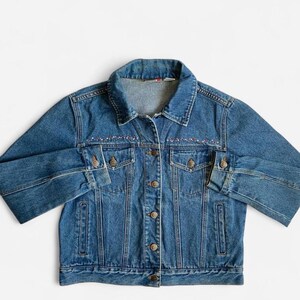 May include: A medium-wash blue denim jacket featuring a classic collar, long sleeves with buttoned cuffs, and a front button closure. The garment includes two chest pockets with buttoned flaps and two vertical side pockets, accented by floral embroidery.
