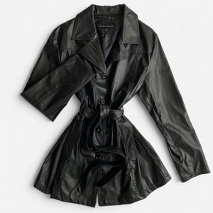 Puede incluir: Un abrigo largo de cuero negro con cinturón en la cintura y cierre de botones. El abrigo tiene un cuello clásico y mangas largas. El abrigo está plano sobre una superficie blanca.