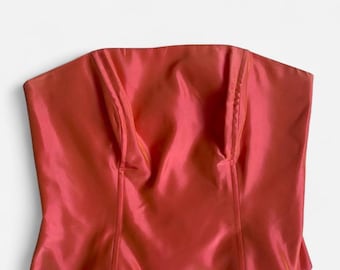 May include: A strapless coral-colored top with a smooth, satin-like finish. The garment features a straight neckline, vertical seam detailing along the front bodice, and a slightly flared hemline, creating a structured and fitted silhouette.