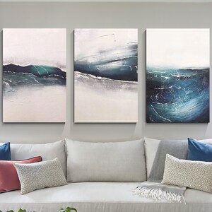 May include: Three abstract paintings depicting a seascape with shades of blue, green, and white. The paintings are hung on a wall above a white sofa with cushions and a throw blanket.