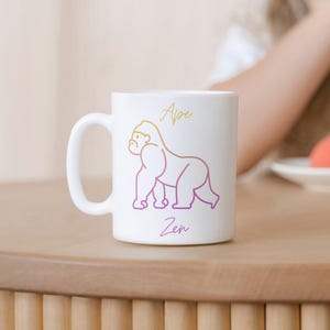 Könnte beinhalten: Weiße Keramiktasse mit einer pinkfarbenen und gelben Umrandung eines Gorillas und dem Text "Ape Zen".