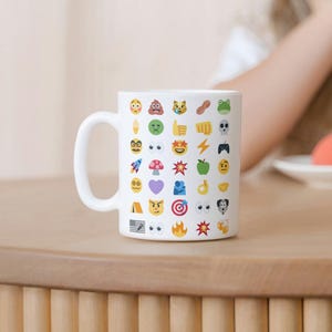 May include: White ceramic mug covered in colorful emoji icons. The mug has a large handle and is sitting on a wooden surface. The emojis include a variety of faces, objects, and symbols, creating a fun and playful design.