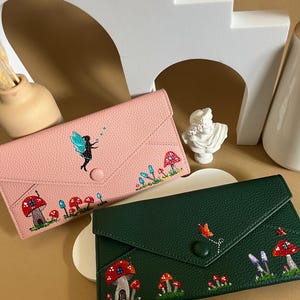 May include: Two leather wallets, one in pink and the other in dark green, with embroidered designs. The pink wallet has a fairy and mushrooms, the green one features mushrooms and butterflies. Both have a button closure.