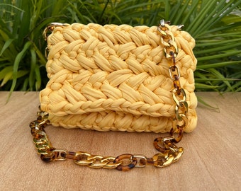 Könnte beinhalten: Eine gelbe gehäkelte Handtasche mit einem goldenen und schildpattfarbenen Kettenriemen.