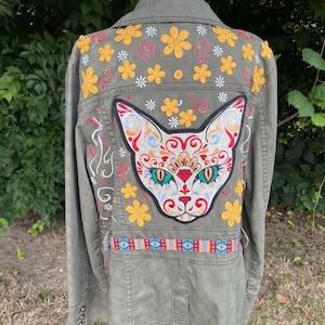 May include: Olive green jacket with embroidered cat face and floral designs. The back features a large cat face patch with colorful details, surrounded by yellow and white flowers. A decorative belt with geometric patterns accents the waist.