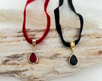 Puede incluir: Dos gargantillas de terciopelo con colgantes de gota de oro. La gargantilla de la izquierda es roja con una piedra preciosa roja, y la gargantilla de la derecha es negra con una piedra preciosa negra.