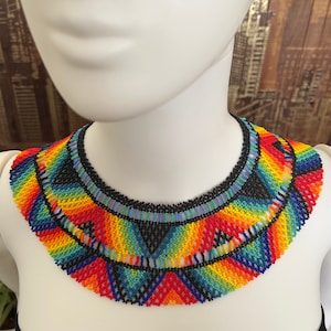 May include: A colorful beaded necklace with a geometric pattern in shades of red, orange, yellow, green, blue, and black. The necklace is made of small, tightly woven beads and has a scalloped edge.