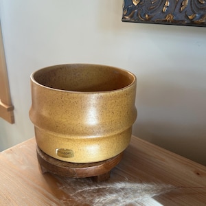 May include: A speckled, yellow-brown ceramic planter with a cylindrical shape and a slightly flared rim. The planter sits on a small, round wooden stand. The label on the planter reads "Haeger."