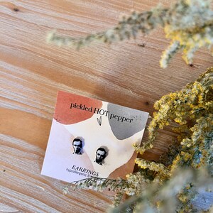 May include: A pair of hypoallergenic stud earrings featuring portraits of Abraham Lincoln. The earrings are displayed on a card with the text "pickled HOT pepper" and "EARRINGS". The background is a light wood surface with dried yellow flowers.