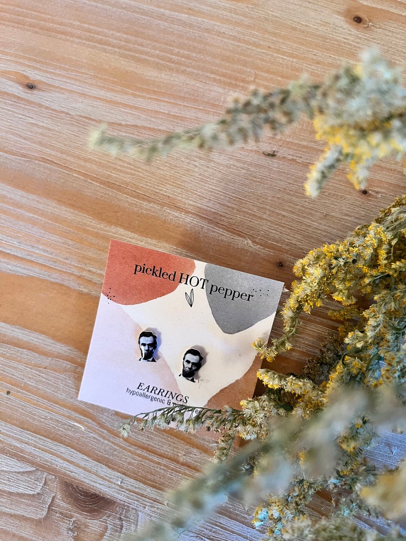May include: A pair of hypoallergenic stud earrings featuring portraits of Abraham Lincoln. The earrings are displayed on a card with the text "pickled HOT pepper" and "EARRINGS". The background is a light wood surface with dried yellow flowers.