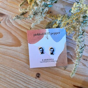 May include: A pair of stud earrings featuring a black and white image of Abraham Lincoln on a pink and blue card. The card reads "pickled HOT pepper" and "EARRINGS hypoallergenic & nickel free". The earrings are displayed on a wooden surface with dried flowers.