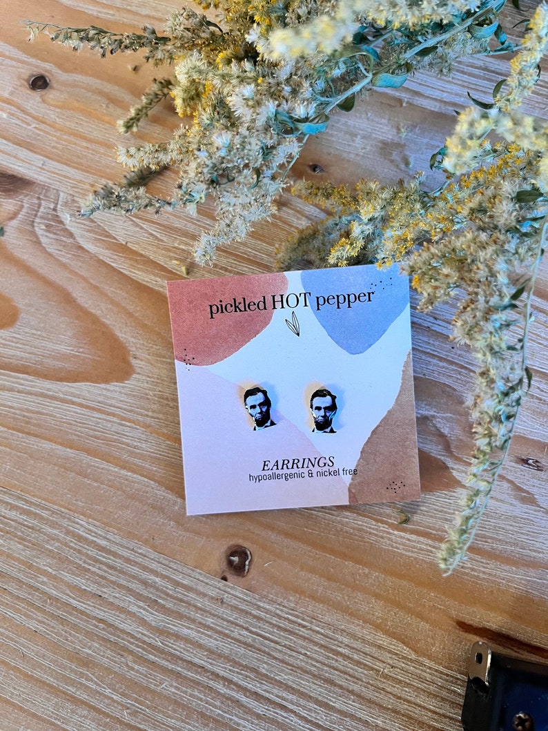 May include: A pair of stud earrings featuring a black and white image of Abraham Lincoln on a pink and blue card. The card reads "pickled HOT pepper" and "EARRINGS hypoallergenic & nickel free". The earrings are displayed on a wooden surface with dried flowers.