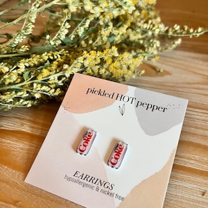 May include: Diet Coke can earrings on a pink and white card with the text "pickled HOT pepper" and "EARRINGS hypoallergenic & nickel free". Dried yellow flowers are in the background.