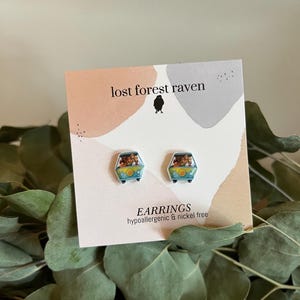 May include: A pair of blue and white stud earrings featuring a cartoon van with passengers. The earrings are hypoallergenic and nickel free. The card reads "lost forest raven" and "EARRINGS hypoallergenic & nickel free".