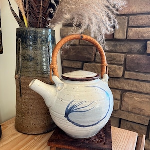 May include: A white ceramic teapot with a brown wooden handle and lid. The teapot has a blue abstract design and sits on a small, dark wooden stand. A tall, textured vase and decorative feathers are in the background.