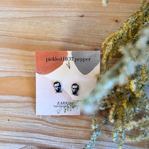 May include: A pair of hypoallergenic earrings featuring a portrait of Abraham Lincoln. The earrings are displayed on a card with the text "pickled HOT pepper" and a watercolor design. The background is a light wood surface.