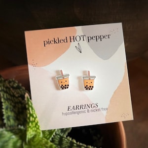 May include: A pair of bubble tea earrings on a card with the text "pickled HOT pepper". The earrings are shaped like bubble tea cups with a smiling face, brown boba, and a straw. The card also says "EARRINGS hypoallergenic & nickel free".