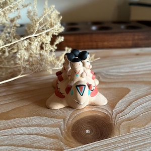 May include: A hand-painted ceramic figurine of a turtle carrying four people on its back. The turtle is painted in shades of brown, red, and blue, with a turquoise gemstone on its head. The people are painted in shades of brown and black, with simple facial features.