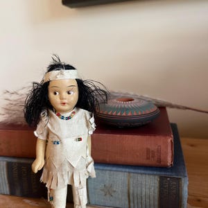 May include: A vintage doll with black hair and a cream-colored outfit, adorned with colorful beads. The doll is standing in front of a stack of books, with a small, decorative ceramic bowl in the background. The doll's outfit has fringe details.