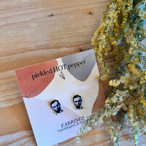 May include: A pair of stud earrings featuring a portrait of Abraham Lincoln. The earrings are displayed on a card with the text "pickled HOT pepper" and "EARRINGS hypoallergenic & nickel free". The background is a wooden surface with dried flowers.