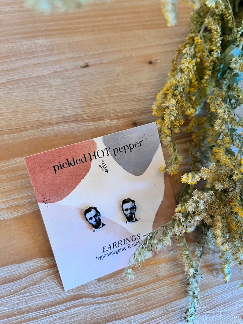 May include: A pair of stud earrings featuring a portrait of Abraham Lincoln. The earrings are displayed on a card with the text "pickled HOT pepper" and "EARRINGS hypoallergenic & nickel free". The background is a wooden surface with dried flowers.
