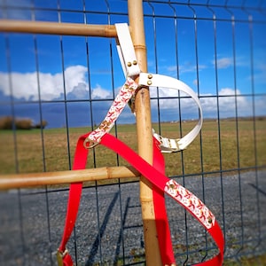 Peut inclure: Deux harnais pour chien, l'un rouge avec un motif floral blanc et l'autre blanc avec des accents dorés, accrochés à un poteau en bois.