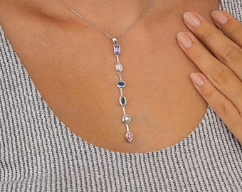 May include: A silver necklace with a vertical pendant featuring a series of colorful gemstones. The gemstones include purple, white, blue, green, and pink, in various shapes. The necklace is worn against a striped shirt.