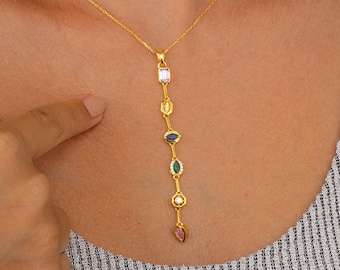May include: A gold-colored necklace with a vertical pendant featuring a series of colorful gemstones. The gemstones include purple, white, blue, green, and pink. The necklace is worn around the neck.