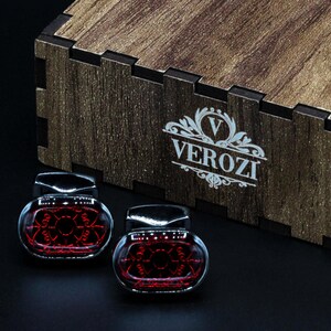 May include: Two glass and chrome cufflinks with a red geometric design are displayed in front of a wooden box. The box has the word "VEROZI" printed on it. The cufflinks are on a black surface.