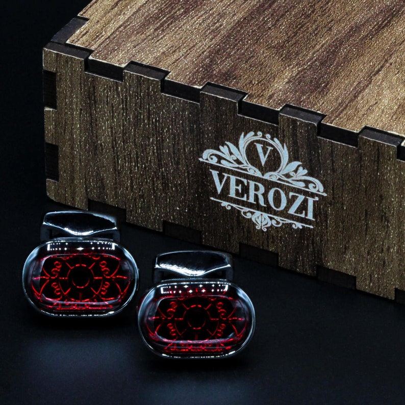 May include: Two glass and chrome cufflinks with a red geometric design are displayed in front of a wooden box. The box has the word "VEROZI" printed on it. The cufflinks are on a black surface.