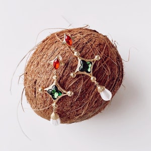 Könnte beinhalten: Goldfarbene Ohrhänger mit roten und grünen Schmuckakzenten. Die Ohrringe haben ein kreuzartiges Design mit einer tropfenförmigen weißen Perle am unteren Ende. Die Ohrringe sind vor einem braunen, strukturierten Hintergrund platziert.