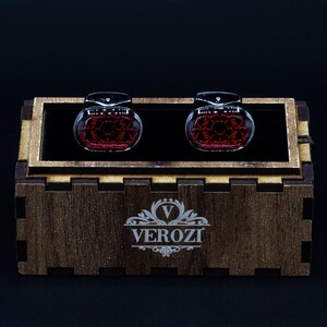 May include: A pair of cufflinks with a red design inside a clear, oval-shaped glass. The cufflinks are displayed in a wooden box with the brand name "VEROZI" engraved on the front. The background is black.