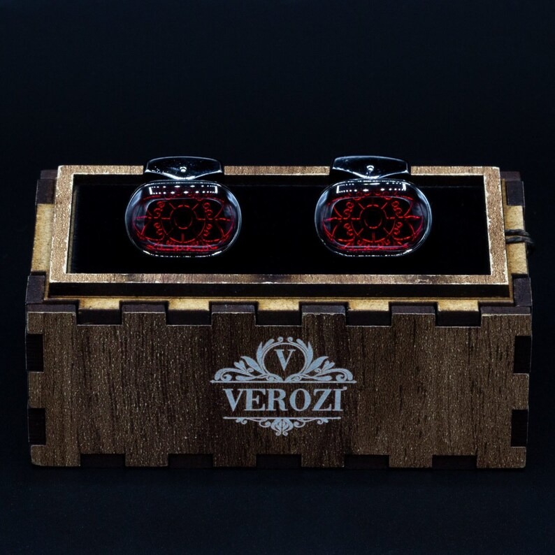 May include: A pair of cufflinks with a red design inside a clear, oval-shaped glass. The cufflinks are displayed in a wooden box with the brand name "VEROZI" engraved on the front. The background is black.