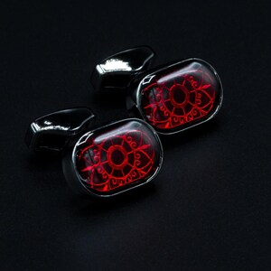 May include: Pair of cufflinks with a red, ornate design set in a silver-tone metal. The cufflinks have a rectangular shape with a glossy finish, and the background is black.