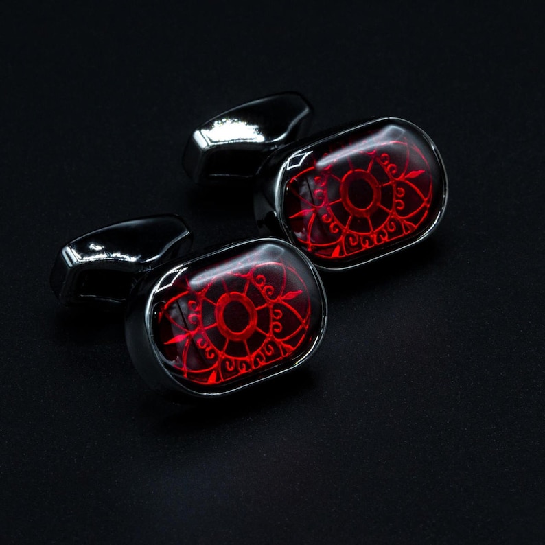May include: Pair of cufflinks with a red, ornate design set in a silver-tone metal. The cufflinks have a rectangular shape with a glossy finish, and the background is black.