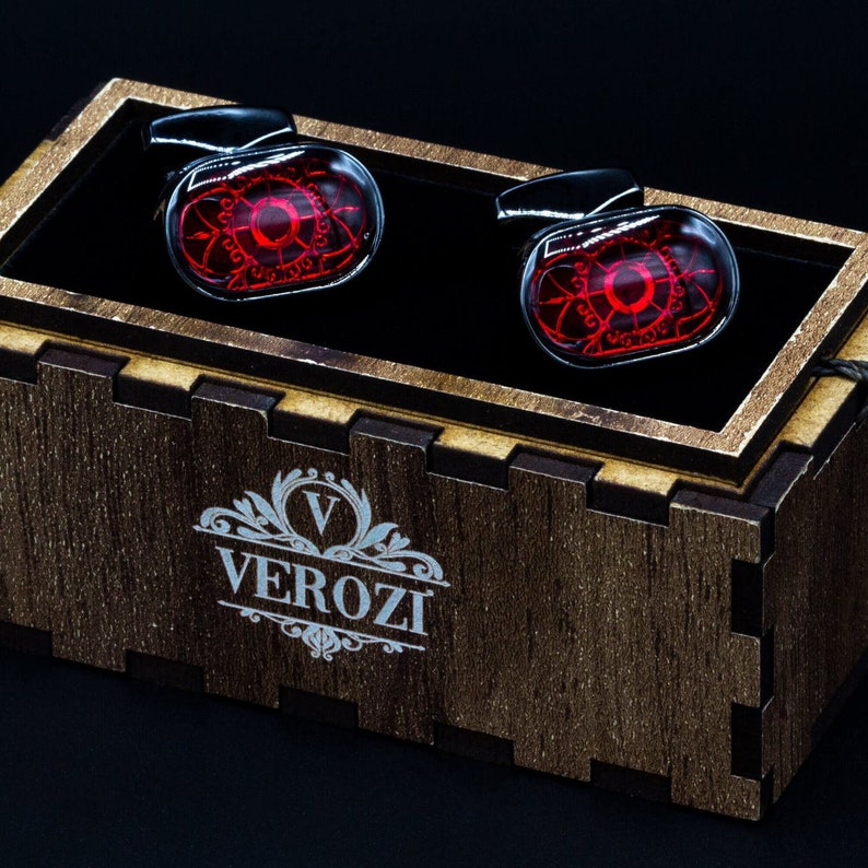 May include: A pair of cufflinks with a red, circular design inside a clear, oval-shaped glass. The cufflinks are set in a wooden box with the word "VEROZI" printed on the front. The cufflinks have a silver-colored backing.